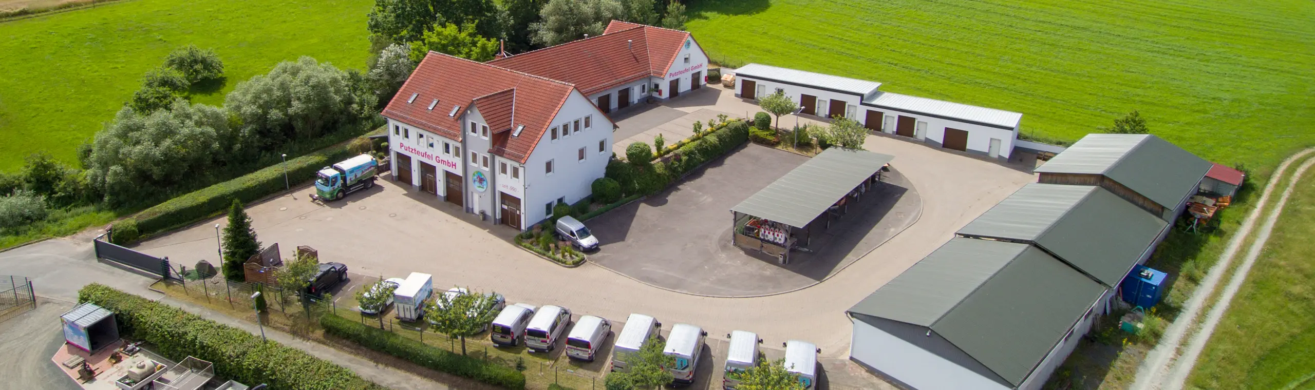 Luftaufnahme des Putzteufel GmbH Firmengeländes mit Hauptgebäude, Hof und Fahrzeugen im Grünen.