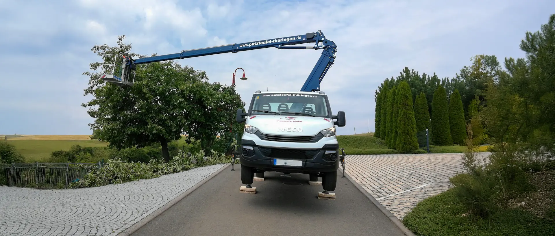Hebebühne mit Arbeiter schneidet Äste von Bäumen entlang einer Straße in gepflegter Gartenanlage.