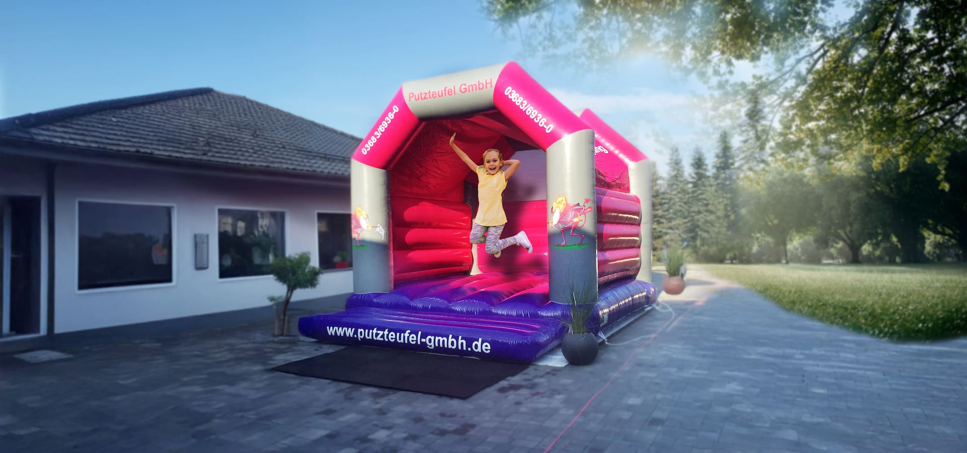 Kind springt fröhlich auf pink-violetter Hüpfburg der Putzteufel GmbH vor Gebäude im Grünen.